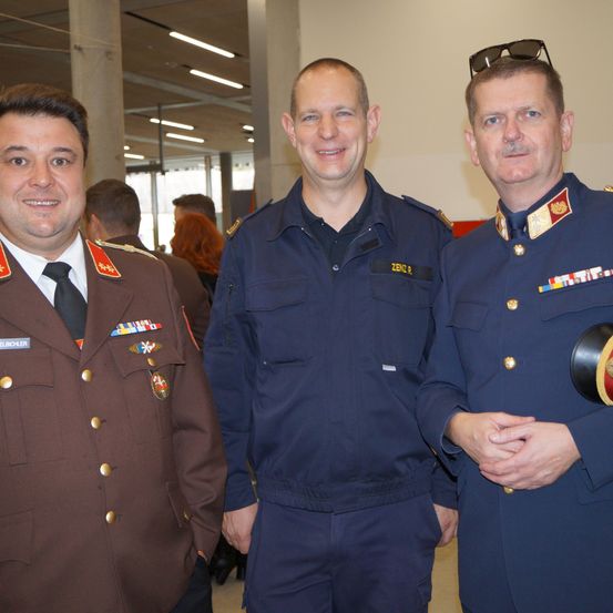 Drei Männer in Militäruniformen, lächelnd und posieren für ein Foto. Ein Mann hat eine Sonnenbrille auf dem Kopf.