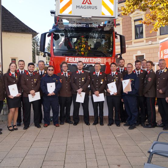 Eine Gruppe von Feuerwehrleuten posiert vor einem roten Feuerwehrauto und hält Urkunden in den Händen. Sie tragen Uniformen mit Medaillen. Gebäude und Bäume sind im Hintergrund.