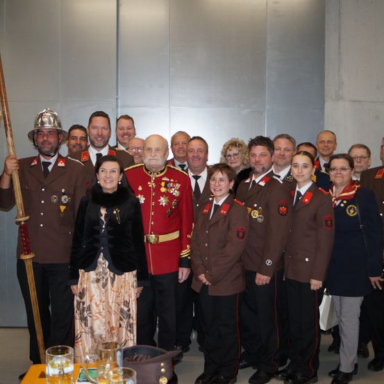 Eine Gruppe von Menschen in Militäruniformen, sowohl Männer als auch Frauen, steht vor einer grauen Wand. Eine Frau in einem schwarzen Kleid steht in der Mitte, während andere Hüte und Brillen tragen.
