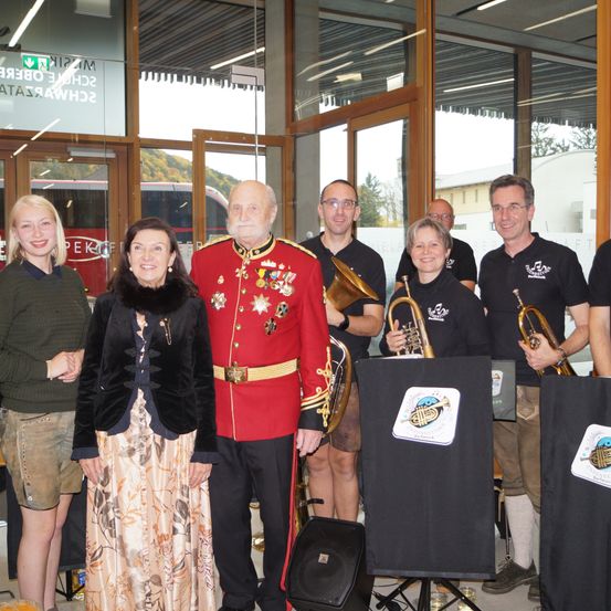Eine Gruppe von Menschen steht in einem Gebäude mit Glaswänden. Ein Mann in einem roten Militäruniform steht in der Mitte. Eine Frau in einem schwarzen Kleid steht neben ihm. Hinter ihnen stehen mehrere Menschen in der Nähe einer Glastür mit einem Logo. Das Gebäude hat eine Glaswand mit Reflexionen von Bäumen und Gebäuden.