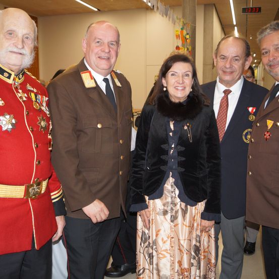 Eine Gruppe formell gekleideter Personen steht in einem Korridor zusammen. Eine Frau in der Mitte lächelt, während andere zuschauen. Sie sind mit Medaillen und Uniformen geschmückt.