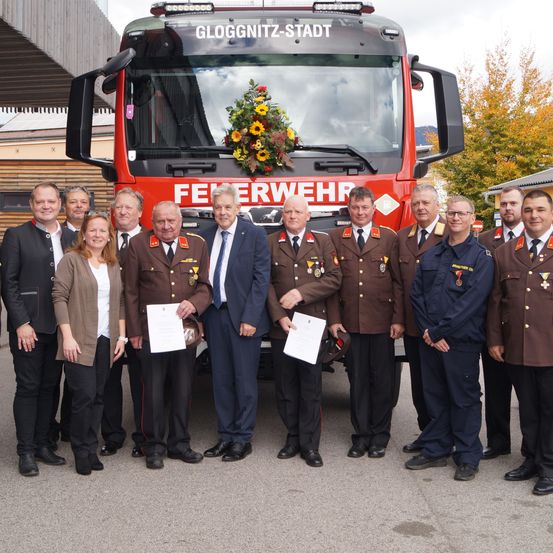 Eine Gruppe von Feuerwehrleuten in Uniform, mit Urkunden, steht vor einem Feuerwehrauto mit Blumen. Dahinter befindet sich ein Gebäude mit einem Baum.