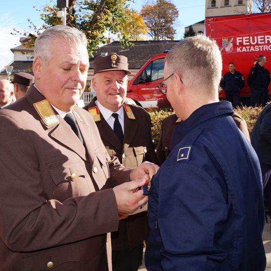 Ein Mann in Uniform befestigt eine Medaille an einem Mann in einem dunkelblauen Jackett. Dahinter steht ein Mann in brauner Uniform, während andere in der Nähe eines roten Fahrzeugs stehen.