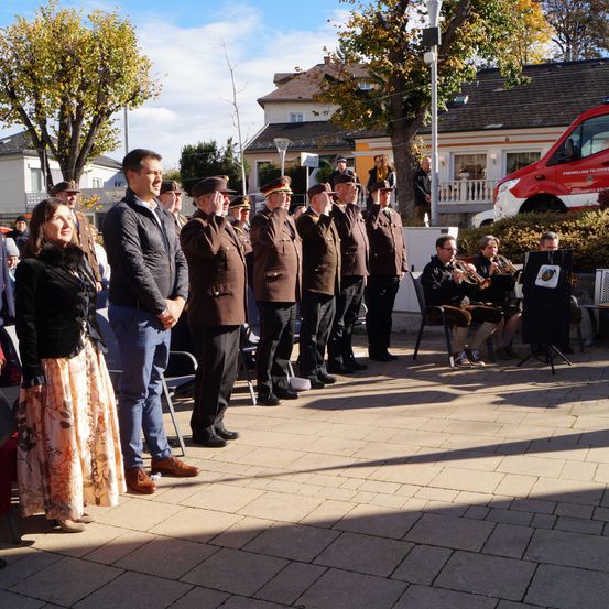 Eine Gruppe uniformierter Personen steht in einer Reihe, wahrscheinlich an einer Zeremonie teilnehmend. Sie sind draußen mit einem roten Fahrzeug, das hinter ihnen geparkt ist. In der Nähe sitzen einige Personen auf Stühlen und spielen Musikinstrumente.