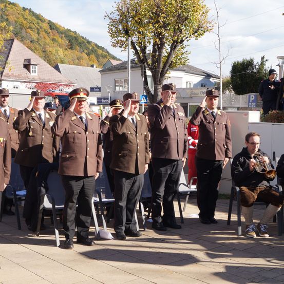 Eine Gruppe von Militäroffizieren steht in einer Reihe, salutiert mit ihren Mützen auf einem öffentlichen Platz. In der Nähe spielt ein Mann Trompete. Im Hintergrund befinden sich Gebäude und Bäume.