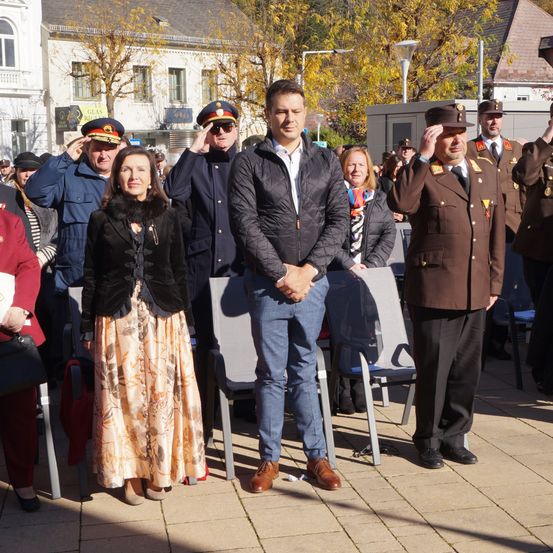 Mehrere Menschen stehen draußen, wahrscheinlich bei einer formellen Veranstaltung. Ein Mann in einer schwarzen Jacke steht mit gefalteten Händen vor sich. Eine Frau in einem Blumenrock steht neben ihm. Hinter ihnen stehen zwei Männer in Militäruniformen, die salutieren.