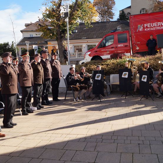 Eine Gruppe von Männern in Uniform steht vor einem Gebäude mit einem roten Lastwagen, ein Mann salutiert, während andere Trompeten und Posaunen spielen.