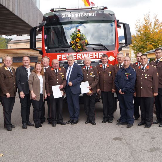 Eine Gruppe von Feuerwehrleuten steht vor einem Feuerwehrauto mit Blumen darauf. Sie halten Papiere in den Händen und tragen Uniformen.