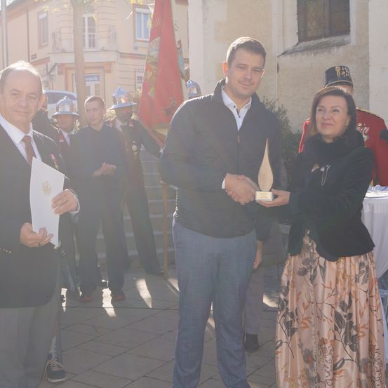 Ein Mann erhält von einer Frau in einem Blumenrock eine Auszeichnung. Hinter ihnen hält ein Mann in Anzug ein Zertifikat. Mehrere Menschen in Uniformen stehen auf den Stufen im Hintergrund.