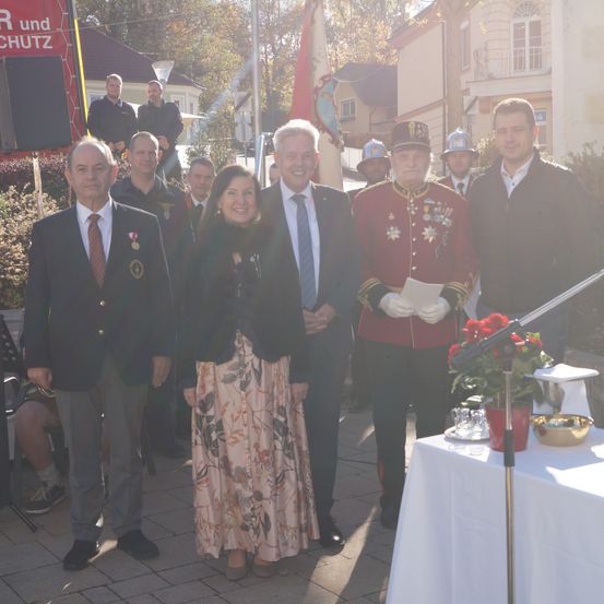 Eine Gruppe von Menschen steht in formeller Kleidung im Freien. Ein Tisch mit Blumen und einem Mikrofon ist aufgebaut. Sie stehen vor einem Gebäude mit Fahnen.