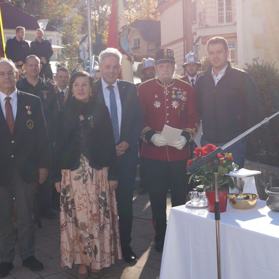Eine Gruppe von Menschen steht vor einem Tisch mit einem Mikrofon, trägt formelle Kleidung und Medaillen. Die Umgebung scheint ein formelles Ereignis oder eine Zeremonie zu sein.