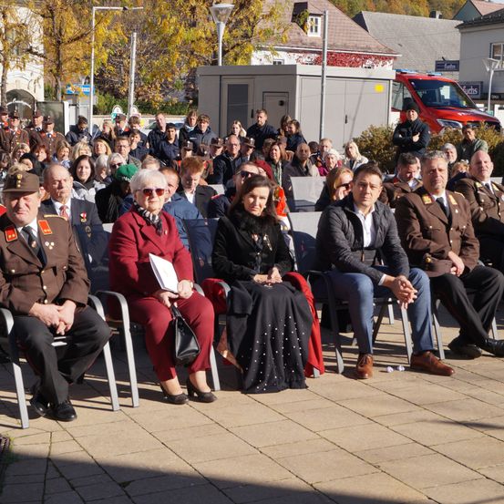 Eine Menschenmenge in formeller Kleidung sitzt in Reihen auf einem gepflasterten Bereich, wahrscheinlich für eine Veranstaltung. In der vorderen Reihe hält eine Frau in einem roten Mantel ein Papier. Mehrere Männer in Militäruniformen sitzen neben ihr. Ein roter Lieferwagen ist in der Nähe geparkt.