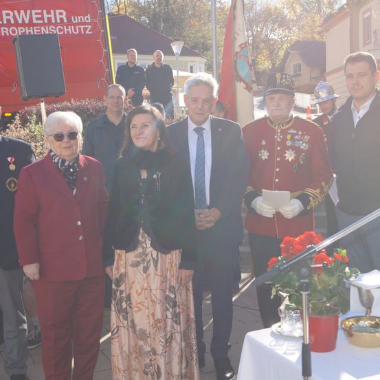 Eine Gruppe von Menschen steht vor einem roten Schild mit einem Banner und Blumen. Ein Mann in Uniform hält ein Papier.