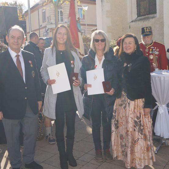 Fünf Menschen stehen auf einem gefliesten Boden und lächeln für ein Foto. Vier von ihnen halten Urkunden. Ein Mann trägt eine Militäruniform. Hinter ihnen steht ein Tisch mit einer Fahne.