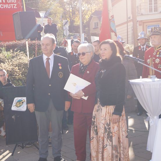 Ein Mann und eine Frau stehen nebeneinander und halten ein Papier in der Hand, wahrscheinlich bei einer öffentlichen Veranstaltung. Hinter ihnen versammeln sich Menschen in Uniformen und Hüten in der Nähe eines Podiums und eines Mikrofons. Bäume und ein Gebäude sind im Hintergrund zu sehen.