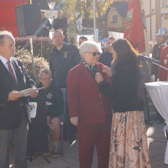Eine offizielle Veranstaltung, bei der eine ältere Frau eine Auszeichnung von einem Mann in einem Anzug erhält. Sie stehen neben einem Mikrofon. Dahinter stehen mehrere Personen, einschließlich eines Mannes, der eine Trompete spielt.