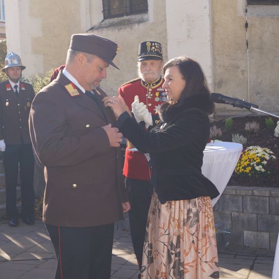 Ein Mann in Uniform wird von einer Frau in einem schwarzen Mantel vor einem Gebäude ausgezeichnet.