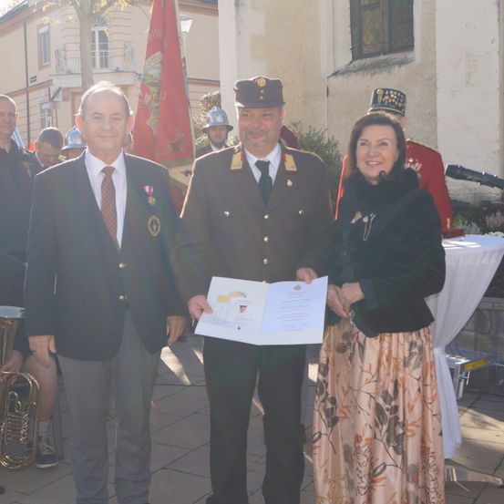 Drei Menschen stehen lächelnd für ein Foto. Ein Mann hält ein Papier, und eine Frau steht neben ihm. Hinter ihnen tragen Menschen Uniformen, und es gibt ein Gebäude mit einer Flagge.