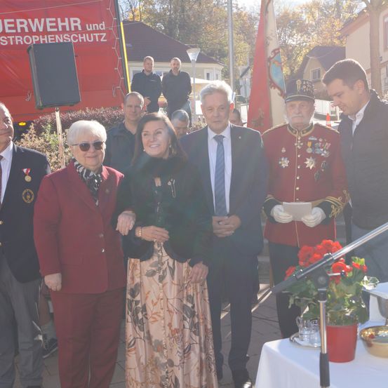 Mehrere Menschen in formeller Kleidung stehen zusammen draußen. Ein Mann in einem roten Uniform hält ein Stück Papier, und eine Frau in einem Blumenkleid lächelt. Dahinter steht ein Tisch mit einer Blumenvase und einem Mikrofon.