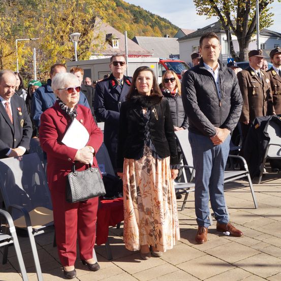 Mehrere Menschen stehen in einer Reihe im Freien, darunter ein Mann im Anzug und zwei Frauen, eine mit einer Handtasche und eine mit einem Schal. Hinter ihnen stehen andere Menschen in Uniformen neben Stühlen und einem Baum.