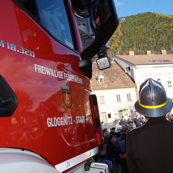 Ein roter Feuerwehrwagen mit 'Freiwillige Feuerwehr' und 'Gloggnitz-Stadt' steht vor einem Gebäude, in dem eine Menschenmenge auf Stühlen sitzt.
