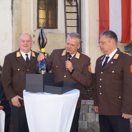 Drei Männer in Militäruniformen stehen um einen Tisch herum und halten eine Trophäe. Der mittlere Mann hält ein Mikrofon und die Trophäe. Ein Banner mit dem Jahr 1914 ist im Hintergrund sichtbar.