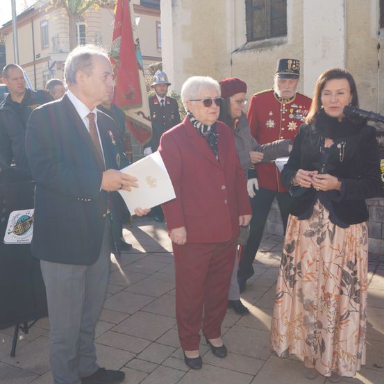 Eine Gruppe formell gekleideter Personen steht in einem Außenbereich. Ein Mann hält ein Stück Papier und eine Frau mit Brille spricht in ein Mikrofon.