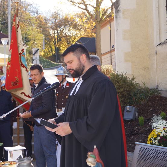 Ein Mann in einer schwarzen Robe liest von einem Tablet neben einem Mikrofon. Hinter ihm steht ein Mann in Helm und Jacke, der zusieht. Im Hintergrund befinden sich eine Fahne und weitere Personen.
