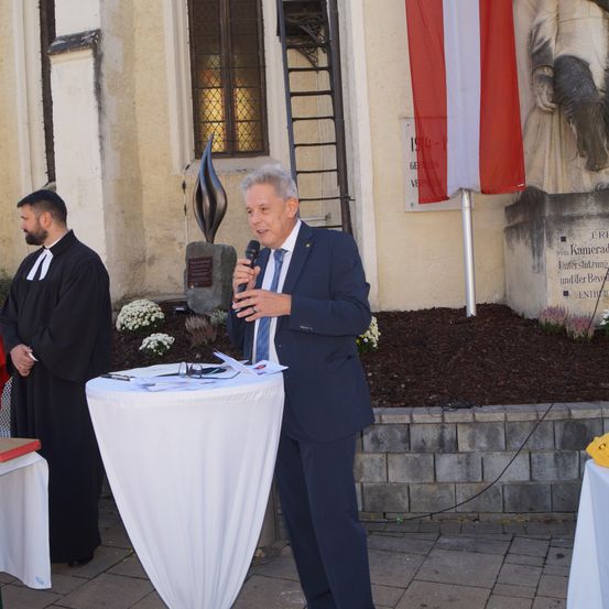 Ein Mann steht an einem Podium und spricht in ein Mikrofon. Neben ihm weht eine Flagge. Hinter ihm steht ein Priester in der Nähe eines Denkmals. Im Hintergrund befinden sich eine Kirche und eine Statue.