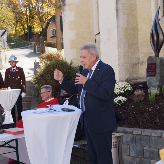 Ein älterer Mann spricht in ein Mikrofon im Freien, mit einer Kirche im Hintergrund. Zwei andere Männer sitzen hinter ihm an einem Tisch, einer trägt einen roten Umhang. Ein Polizeibeamter steht hinter dem Tisch, mit einer Flammenskulptur auf der rechten Seite.