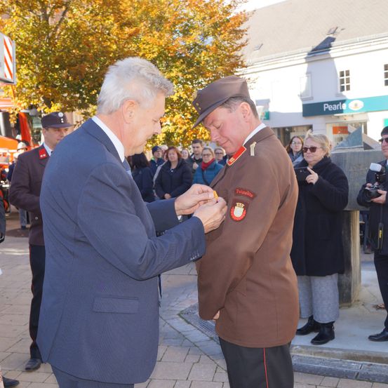 Ein Mann in einem Anzug befestigt eine Medaille an einem Militäroffizier. Sie stehen vor einer Menge, mit Menschen hinter ihnen, die zusehen. Bäume und ein Gebäude sind im Hintergrund.