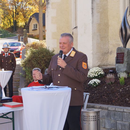 Ein Mann in Uniform spricht bei einer Outdoor-Veranstaltung, er steht bei einem Rednerpult, mit einem Denkmal dahinter und zwei Personen im Hintergrund.