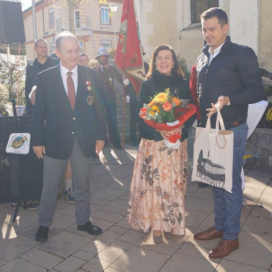 Ein Mann und eine Frau halten Blumen vor einem lächelnden Mann, wahrscheinlich bei einer Gemeindeveranstaltung. Dahinter sind Leute, eine Fahne und ein Gebäude.