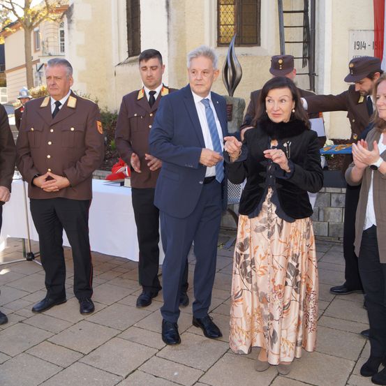 Mehrere uniformierte Männer und Frauen stehen vor einem Gebäude, möglicherweise bei einer Zeremonie. Ein Mann in Anzug und Krawatte hält die Hand einer Frau in einem Kleid.