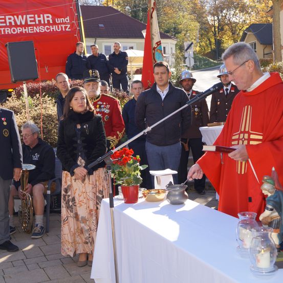 Eine religiöse Zeremonie findet im Freien statt. Ein Mann in einem roten Gewand liest aus einem Buch. Eine Frau in einem schwarzen Kleid steht neben einem Tisch mit Blumen und Kerzen. Mehrere Personen in Uniform stehen hinter einem Banner.