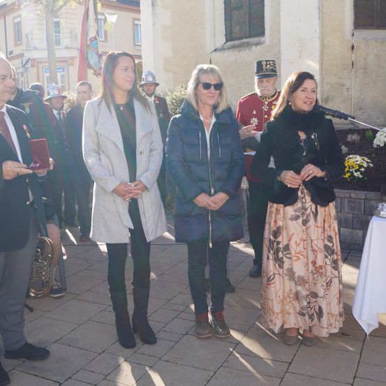 Eine Gruppe von Personen, darunter Frauen und Männer, steht im Freien in einer Reihe. Eine Frau hält ein Mikrofon und lächelt, während andere hinter ihr stehen. Dahinter befinden sich Pflanzen, ein Gebäude und ein Fahnenmast.