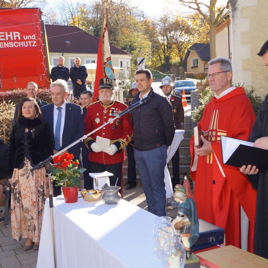 Eine Gruppe von Menschen in formeller Kleidung, die hinter einem Tisch mit Blumen und einem Mikrofon vor einer Kirche stehen.