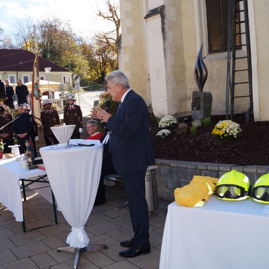 Ein Mann spricht bei einer Outdoor-Veranstaltung vor einem Gebäude, mit uniformierten Feuerwehrleuten im Hintergrund. Tische mit gelben Helmen und Schutzausrüstung sind aufgestellt.