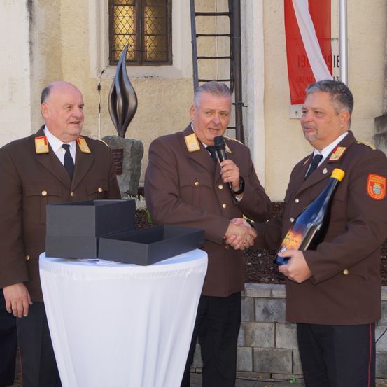Drei Männer in Uniform stehen vor einem Tisch mit Kisten, einer hält eine Flasche und ein Mikrofon, ein anderer schüttelt Hände. Dahinter steht ein Gebäude mit einer Flagge und einer Skulptur.