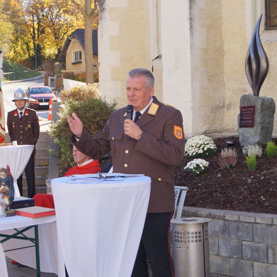 Ein Mann in Uniform steht an einem Podium und hält eine Rede. Hinter ihm eine Statue und ein Gebäude. Ein Mann in einem roten Mantel hört zu. Bäume und ein Auto im Hintergrund.