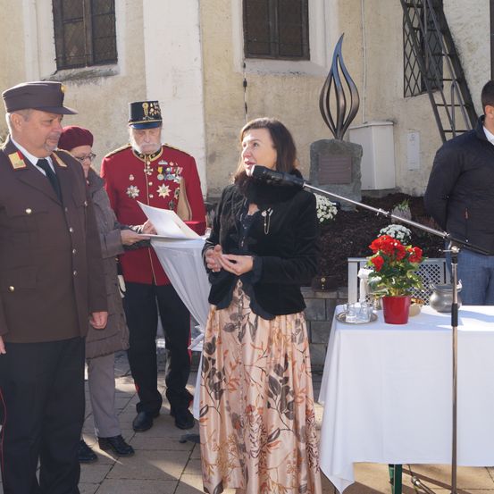 Eine Frau spricht in ein Mikrofon, während zwei Männer in Militäruniformen und ein Mann in einem Anzug hinter ihr stehen. Ein Tisch mit Blumen und einer Vase befindet sich in der Nähe.