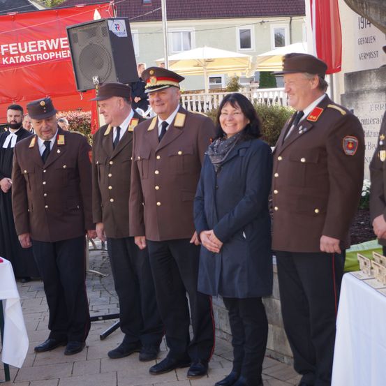 Fünf Personen in formellen Uniformen, darunter eine Frau, stehen vor einem Denkmal. Dahinter befindet sich ein roter Banner, Gebäude und ein Lautsprecher.