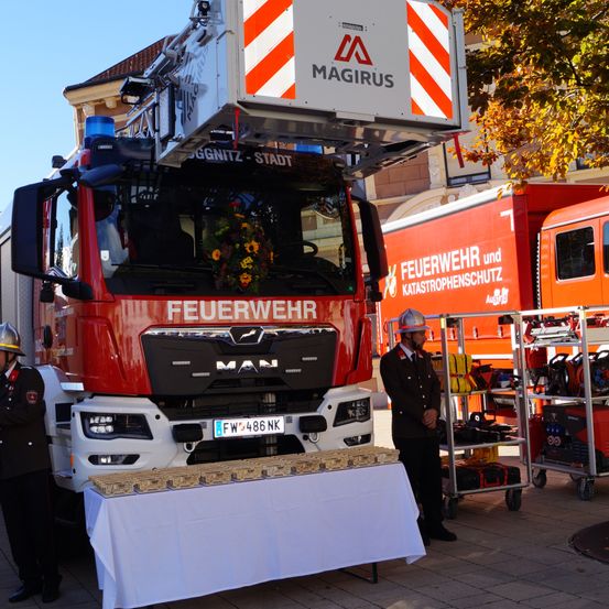 Ein roter Feuerwehrwagen mit Magirus obenauf und einem Blumenstrauß davor, geparkt mit einem Tisch und einem Feuerwehrmann in der Nähe.