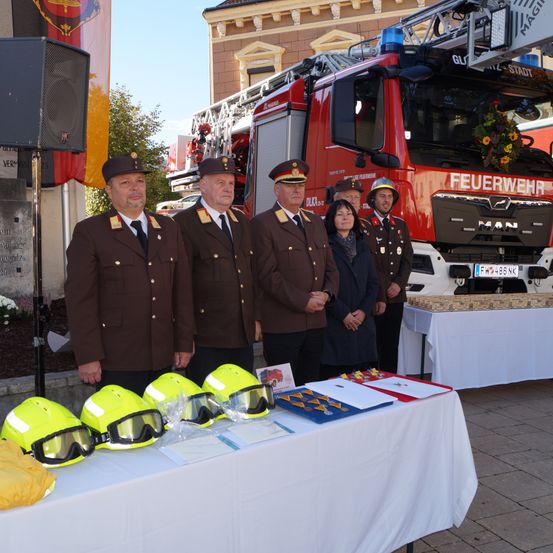 Fünf Personen in Uniform stehen vor einem Feuerwehrauto mit Blumen darauf. Sie stehen neben einem Tisch mit Helmen und Auszeichnungen.