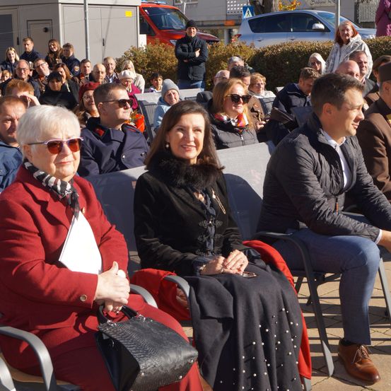 Eine Gruppe von Menschen sitzt in Stühlen, einige mit Brillen und Mänteln, eine Frau in einem roten Mantel hält eine schwarze Handtasche. Hinter ihnen lächelt ein Mann in einem braunen Mantel. Autos und Gebäude sind im Hintergrund.