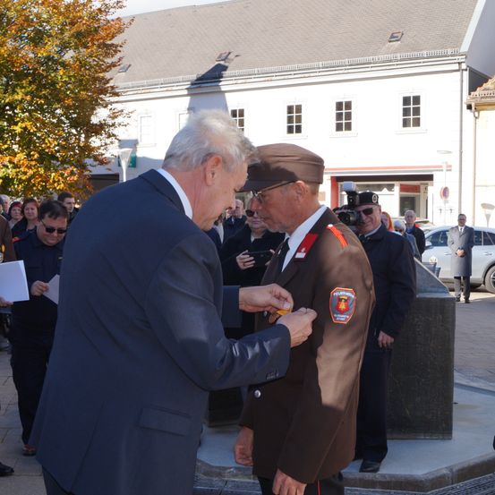 Eine Veranstaltung im Freien, bei der ein älterer Mann in einem Anzug eine Medaille an einen anderen Mann in Uniform anheftet. Hinter ihnen beobachtet eine Menschenmenge, einige halten Kameras. Ein Gebäude mit vielen Fenstern und einem Baum ist im Hintergrund.