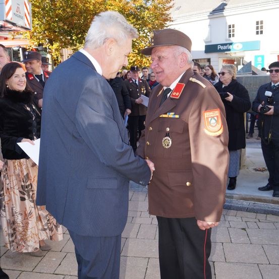 Zwei Männer, einer in Anzug und der andere in Uniform, schütteln sich die Hände bei einer öffentlichen Zeremonie. Dahinter beobachten mehrere Menschen, darunter ein Fotograf. Im Hintergrund sind ein Gebäude und Bäume zu sehen.