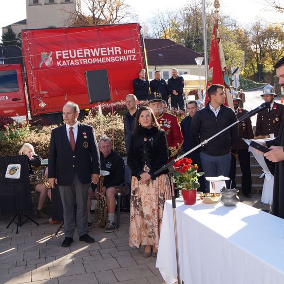 Eine Gruppe von Menschen steht vor einem roten Feuerwehrfahrzeug. Ein Mann spricht in ein Mikrofon, während eine Frau und andere zuhören. Dahinter befinden sich Fahnen und ein Tisch mit Blumen.