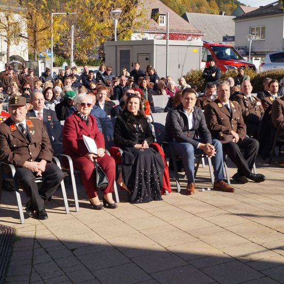 Eine Versammlung von Menschen in Uniformen und formeller Kleidung sitzt auf Stühlen im Freien. Gebäude und Fahrzeuge sind im Hintergrund zu sehen.