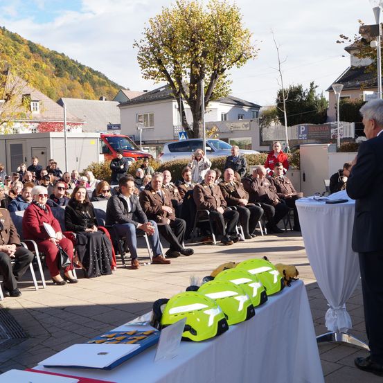 Ein Mann spricht an einem Podium, während eine Menschenmenge zuhört, die auf Stühlen sitzt und steht. Ein Tisch mit Helmen und anderen Gegenständen befindet sich in der Nähe.